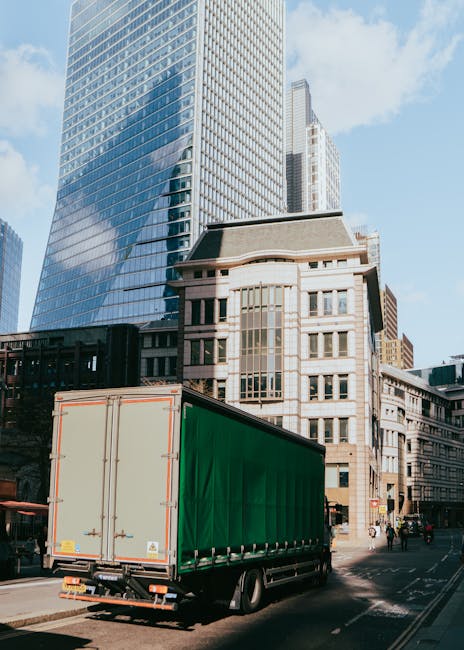 A large green cargo truck is parked on a city street during daylight hours, with the rear doors closed, ready for loading or unloading. The truck is positioned near modern and historic buildings, incl