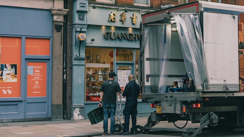 A large green cargo truck is parked on a city street during daylight hours, with the rear doors closed, ready for loading or unloading. The truck is positioned near modern and historic buildings, incl