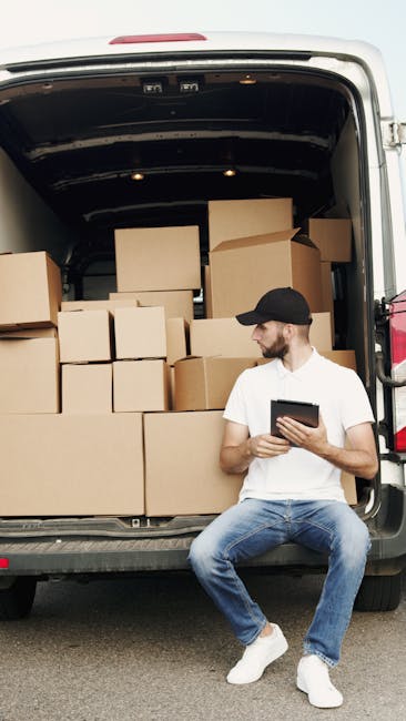 A man wearing a black cap, white t-shirt, and blue jeans is seated on the rear bumper of a van with its back doors open. Inside the van, there are numerous cardboard boxes of various sizes stacked to