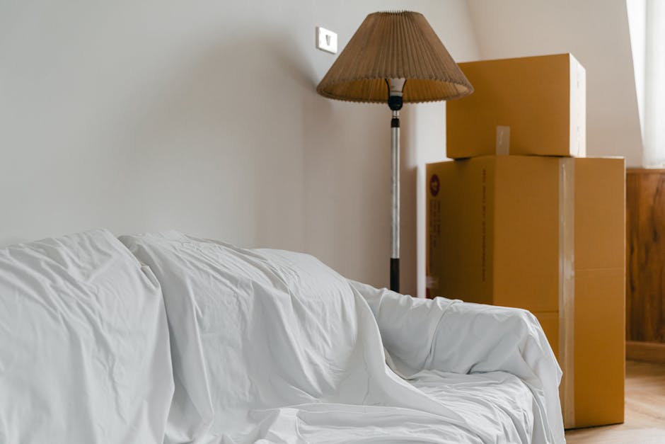 Inside a room during a home relocation, there are three large cardboard boxes stacked in the corner, with one box slightly smaller and placed on top of two larger boxes. To the right of the boxes, the